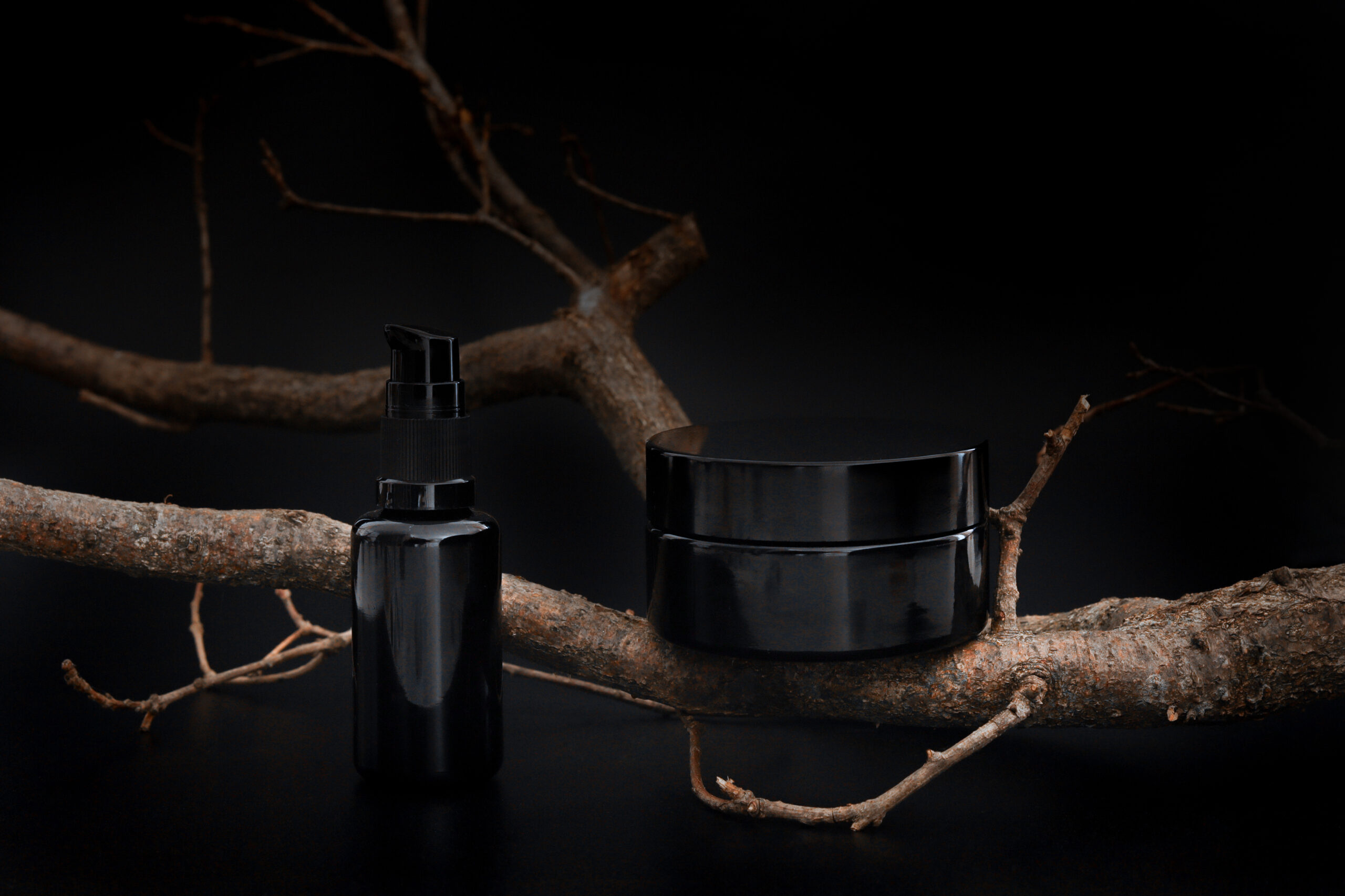 Black unbranded cosmetic cream jar and serum with tree branch. Skin care product presentation on the black background. Elegant mockup. Skincare, beauty and spa.